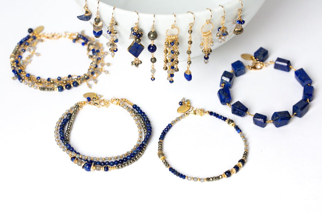 Handcrafted Anne Vaughan Designs Blue Moon collection jewelry featuring deep blue lapis lazuli, pyrite, and labradorite dangle earrings displayed on a ceramic bowl beside layered bead bracelets.