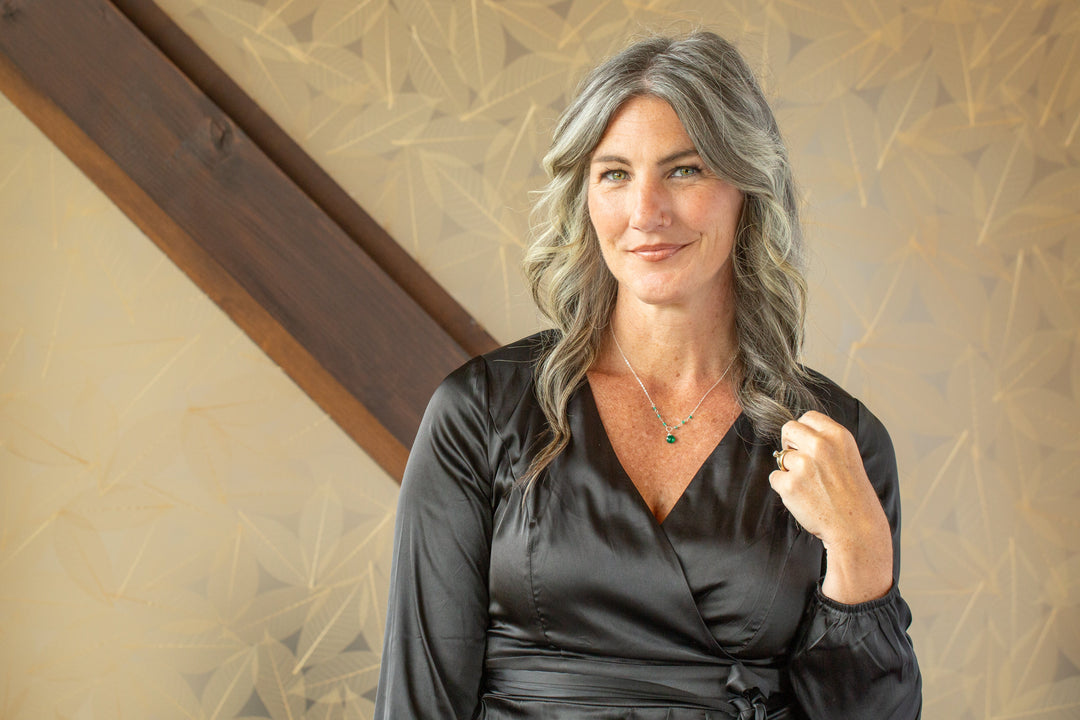 Woman with wavy gray hair wearing a black satin wrap top and an Anne Vaughan Designs green gemstone birthstone necklace.